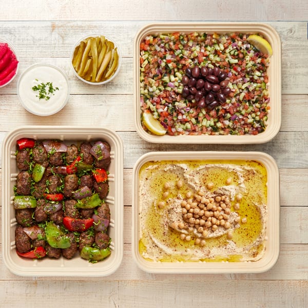 Mediterranean feast with kofta, hummus, salad, pickles, and garlic sauce on a wooden table.