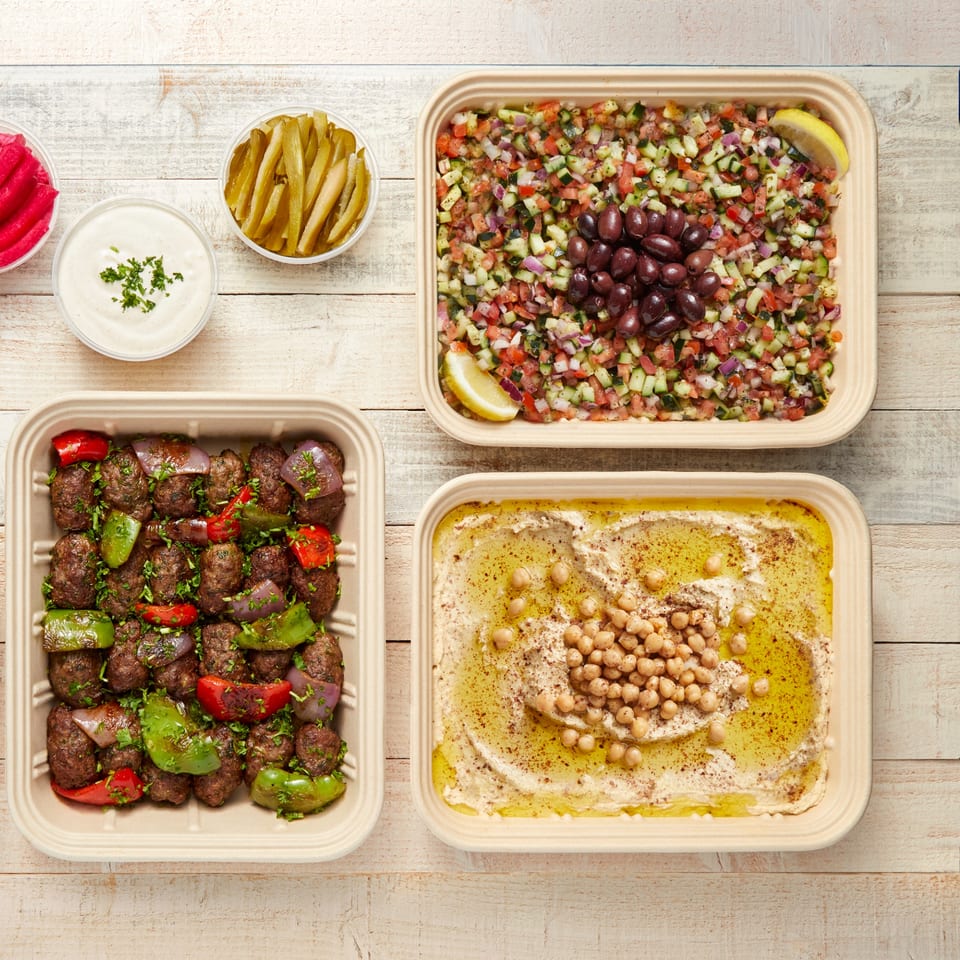 Mediterranean feast with kofta, hummus, salad, pickles, and garlic sauce on a wooden table.