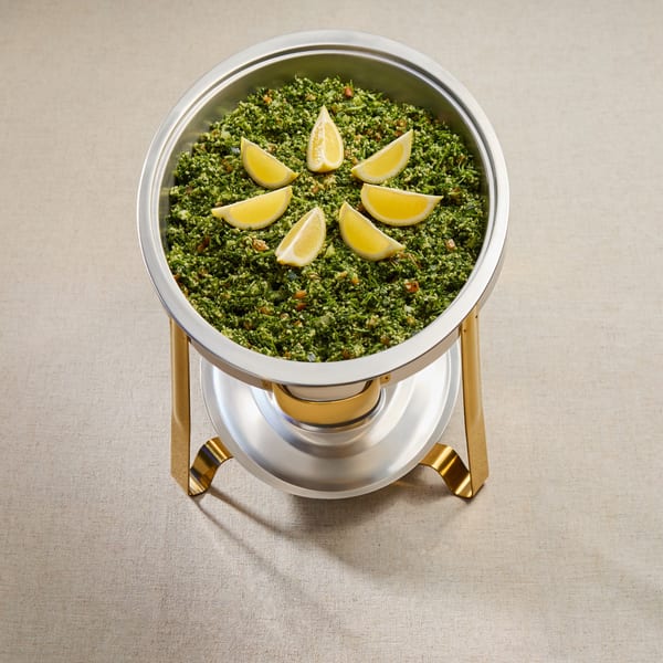 A large silver chafing dish filled with green tabbouleh salad, garnished with lemon wedges.
