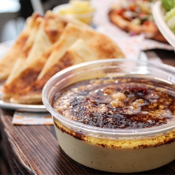 A container of hummus topped with chickpeas and olive oil, with pita bread in the background.