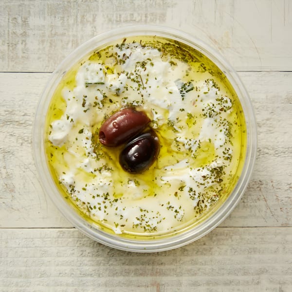 A clear plastic container of labneh with olive oil, herbs, and two olives on a white wooden surface.