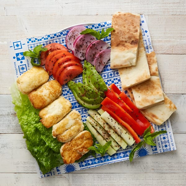 Mediterranean platter with grilled halloumi, sliced tomatoes, red onions, avocado, bell peppers, cucumbers, and pita bread.