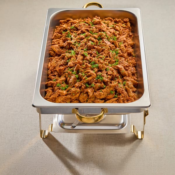 A large metal chafing dish filled with seasoned chicken shawarma, garnished with fresh parsley.